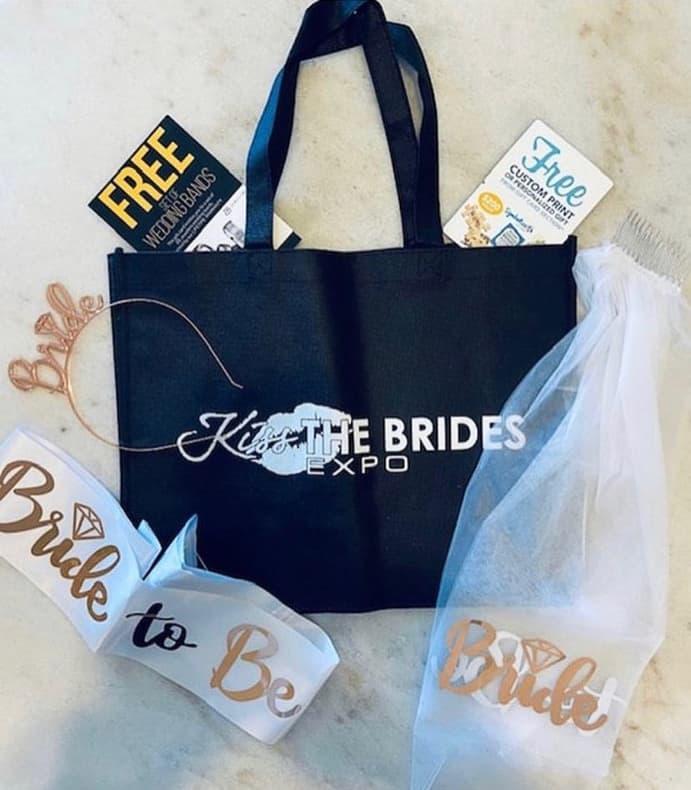 A black tote bag labeled Kiss the Brides Expo with a white veil, a Bride to Be sash, and promotional flyers on a white surface.