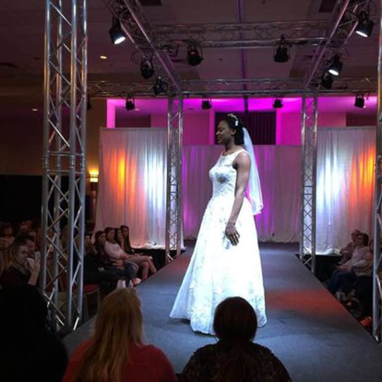 A model in a white wedding dress and veil walks down a runway at a fashion show, with a backdrop of white and pink lighting.