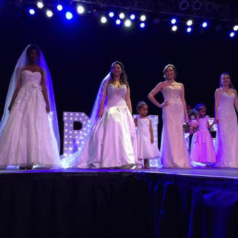 Five models in wedding dresses, accompanied by three child models in white dresses, stand on a well-lit stage at a wedding show.