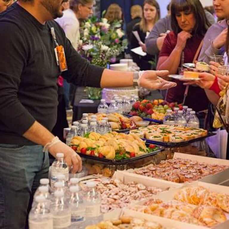 A man serves food to attendees from a table filled with various dishes and bottled water at an event.