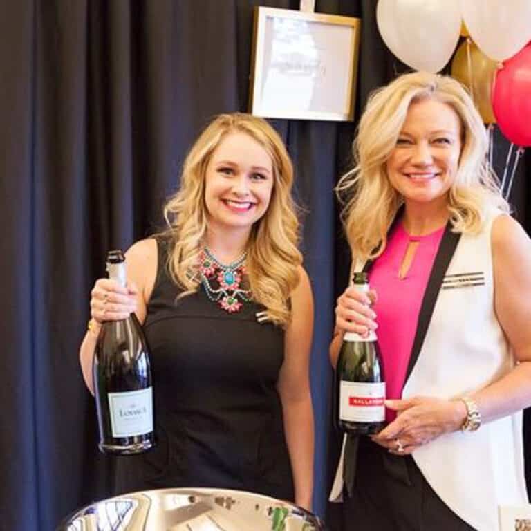 Two women holding champagne bottles stand in front of black curtains with balloons in the background.