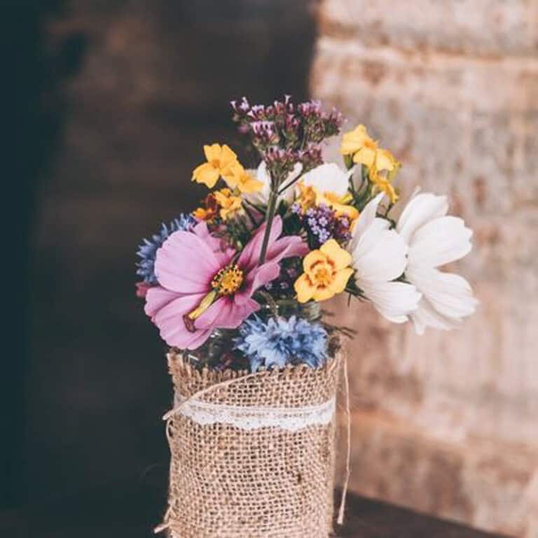 A small bouquet of colorful flowers in a burlap-wrapped vase with lace trim, set against a blurred background.