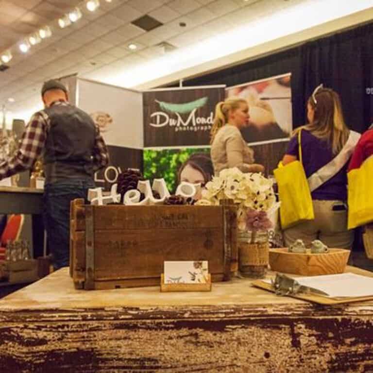 A booth at Kiss the Brides Expo with a wood table holding decorations and business cards, and three people in the background.