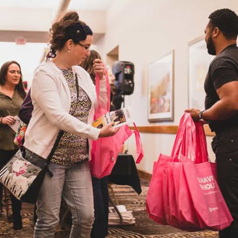 People at a wedding show receiving pink tote bags from a man. One woman is holding a pamphlet and reaching for a bag.