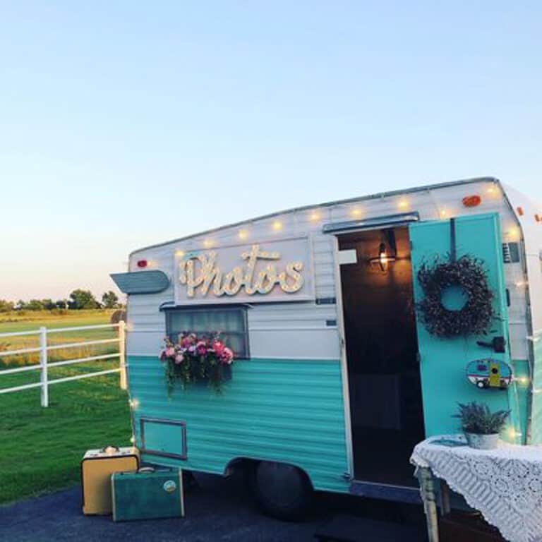 A vintage teal and white camper with Photos sign, decorated with flowers and lights, sits in an open outdoor setting near a white fence.